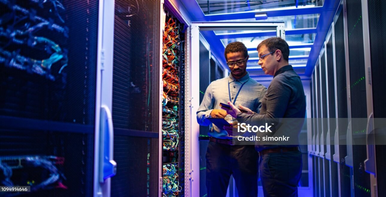 Two male IT engineers using digital tablet and smart phone in server room.