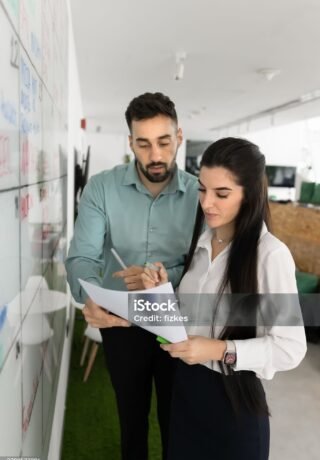 Two business colleagues analyzing paper legal document at white scrum board in office space, reading file together, reviewing contract, agreement text, financial report. Vertical shot