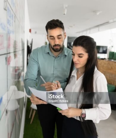 Two business colleagues analyzing paper legal document at white scrum board in office space, reading file together, reviewing contract, agreement text, financial report. Vertical shot